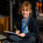 A child with a book and pencil sat on the floor looking at the camera.