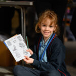A child with a book and pencil sat on the floor looking at the camera and showing their work.