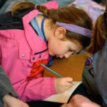 A child in a pink coat writing in their note book.
