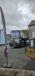 A black bus with the word 'pulse' written on the top deck. It stands in a carpark with a flag/ banner in front of it.