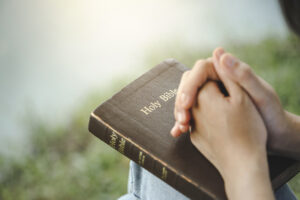 Someone's hands held together resting on a brown book which reads 'Holy Bible'. 